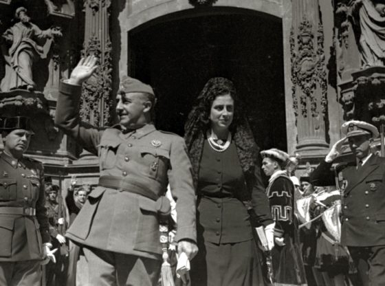 Black and white photo of Spanish dictator Franco and wife waving to supporters outside ornate stone doorway